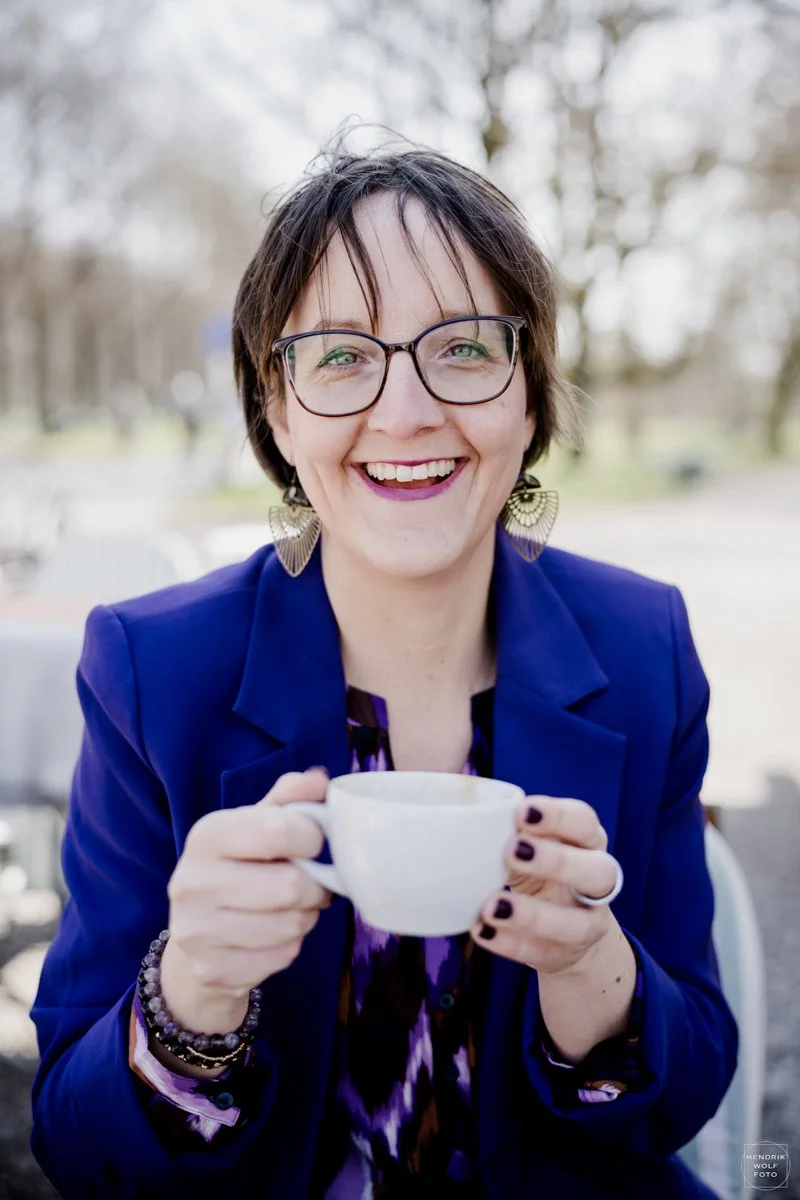 A woman with short brown hair and glasses, smiling and holding a white coffee cup outdoors, wearing a purple and blue outfit.