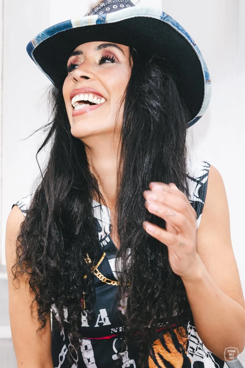 A woman with long black curly hair, wearing a wide-brimmed hat with a blue and white pattern, laughing and looking up, dressed in a sleeveless top with chains and print design.