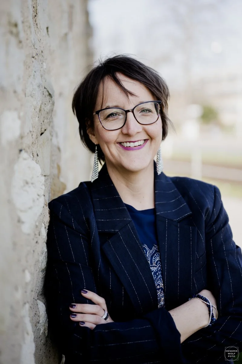A smiling woman with short dark hair, glasses, and earrings, standing outdoors against a textured stone wall, wearing a dark pinstripe blazer and navy shirt.