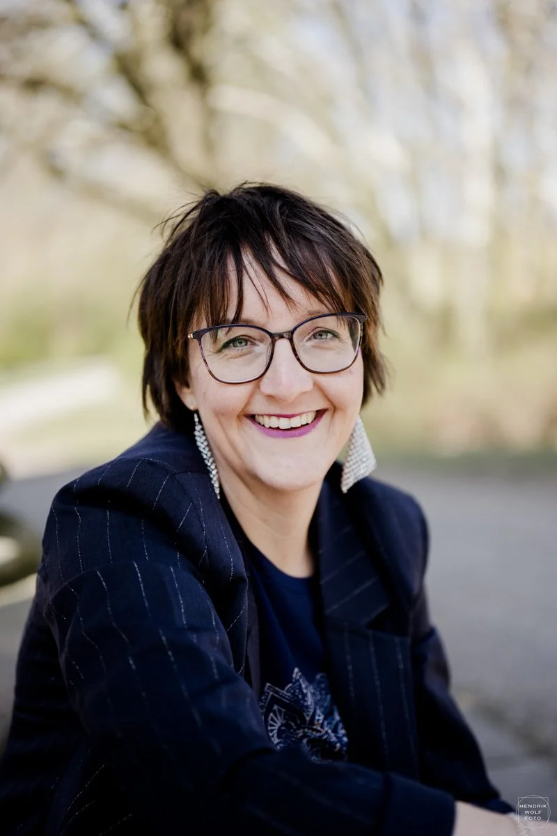 A smiling woman with short dark hair, wearing glasses, earrings, a dark striped blazer, and a graphic shirt, sitting outdoors with trees in the background.