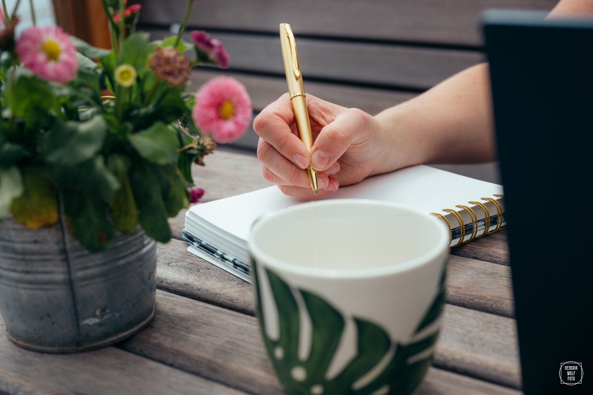 A person's hand writing in a notebook with a gold pen, on a wooden table next to a potted plant, a ceramic cup, and a laptop, with a blurred outdoor background.