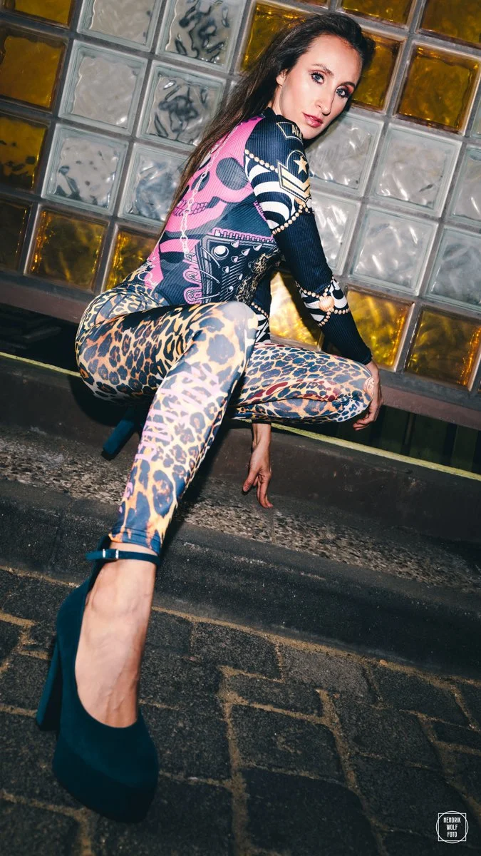 A woman with long dark hair sitting on a curb in front of a wall made of glass bricks with yellow and gray tones, wearing leopard print leggings, a colorful patterned top, and teal high heels.