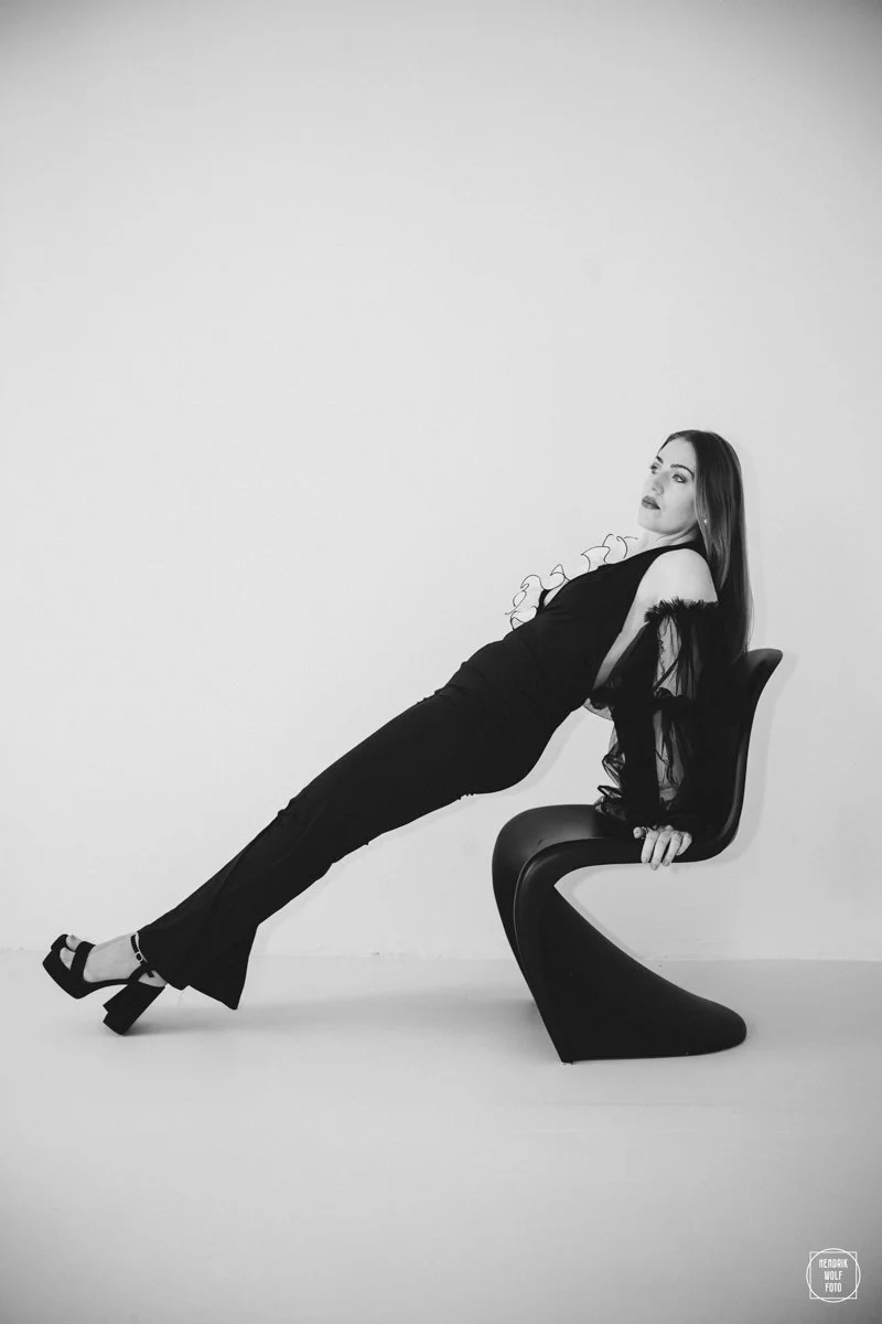 A woman in a black outfit wearing high heels, reclining backwards on a modern black chair against a plain white background.