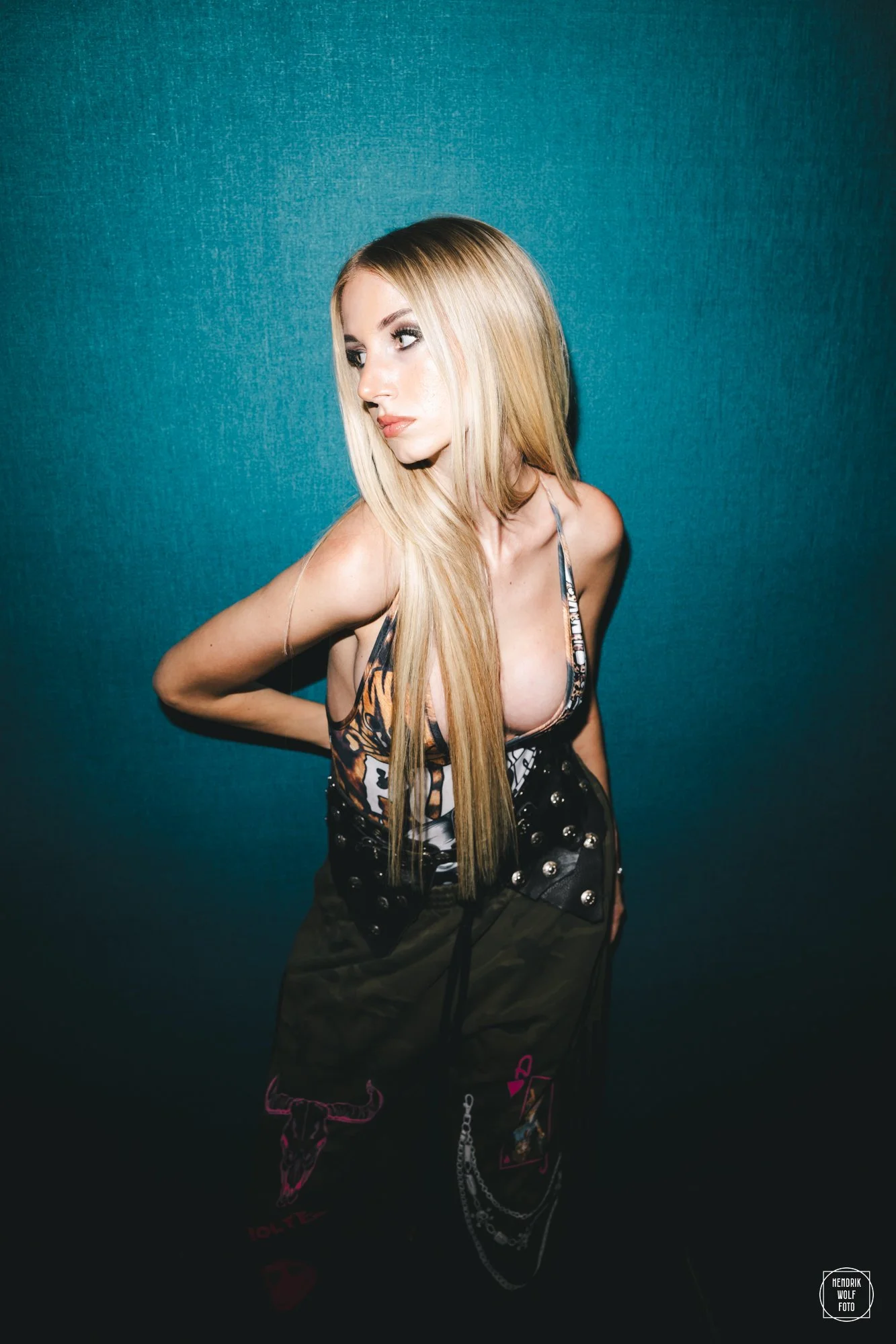A blonde woman with long hair, wearing a black leather top with silver studs and dark pants with chains and pink accents, posing against a teal wall with her left arm bent and her hand on her hip, looking to the left with a serious expression.