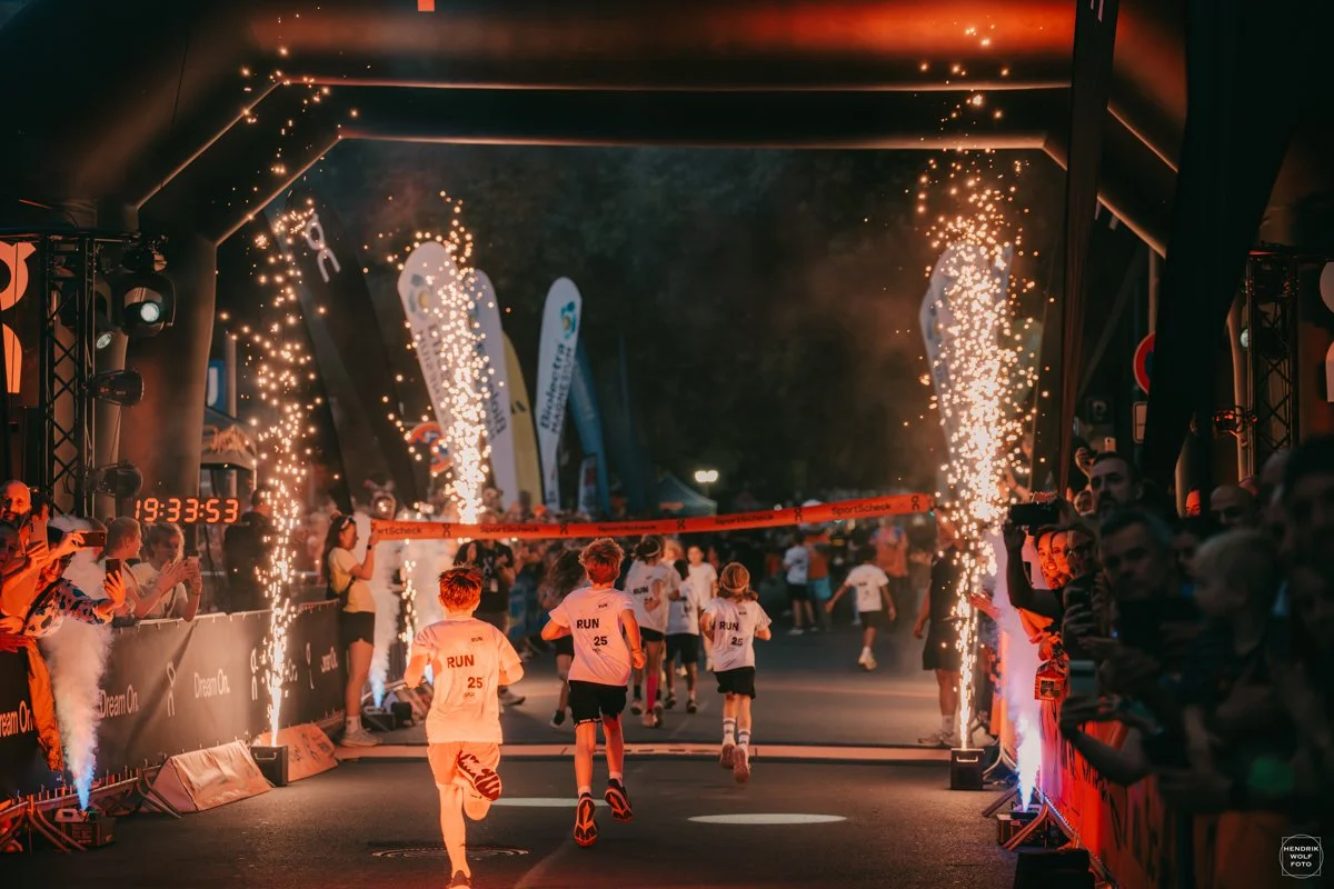 Children running through a nighttime finish line at a race, with sparklers and fireworks lighting the scene, spectators cheering on the sides.