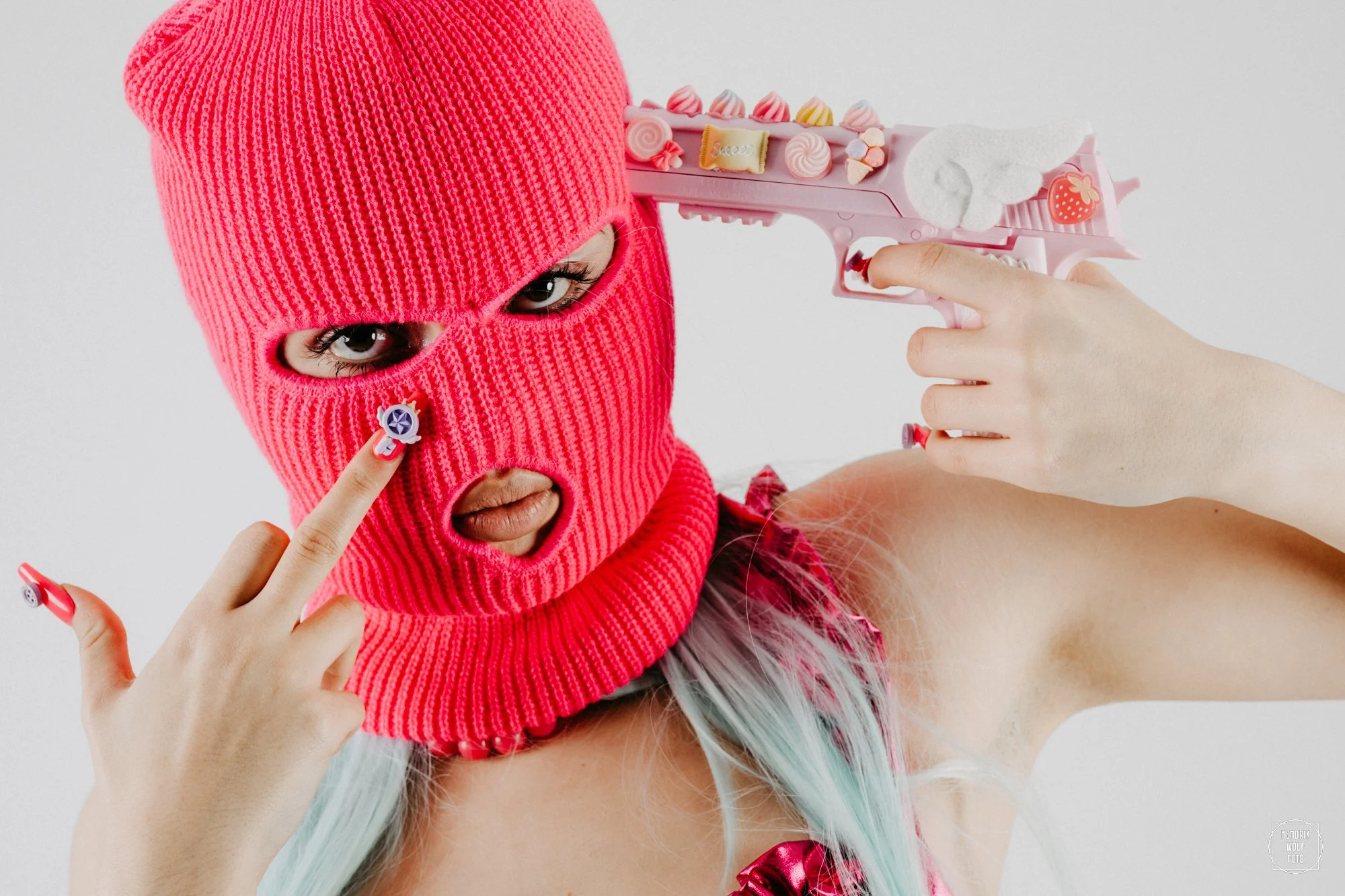 Person wearing a pink ski mask with cut-out eye holes, holding a pink toy gun decorated with candies and sweets, pointing it at their temple.