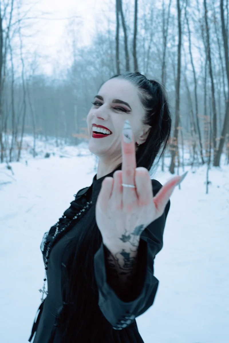 A woman with dark hair, red lipstick, and tattoos on her hand, smiling and showing her middle finger to the camera in a snowy, wooded outdoor setting.
