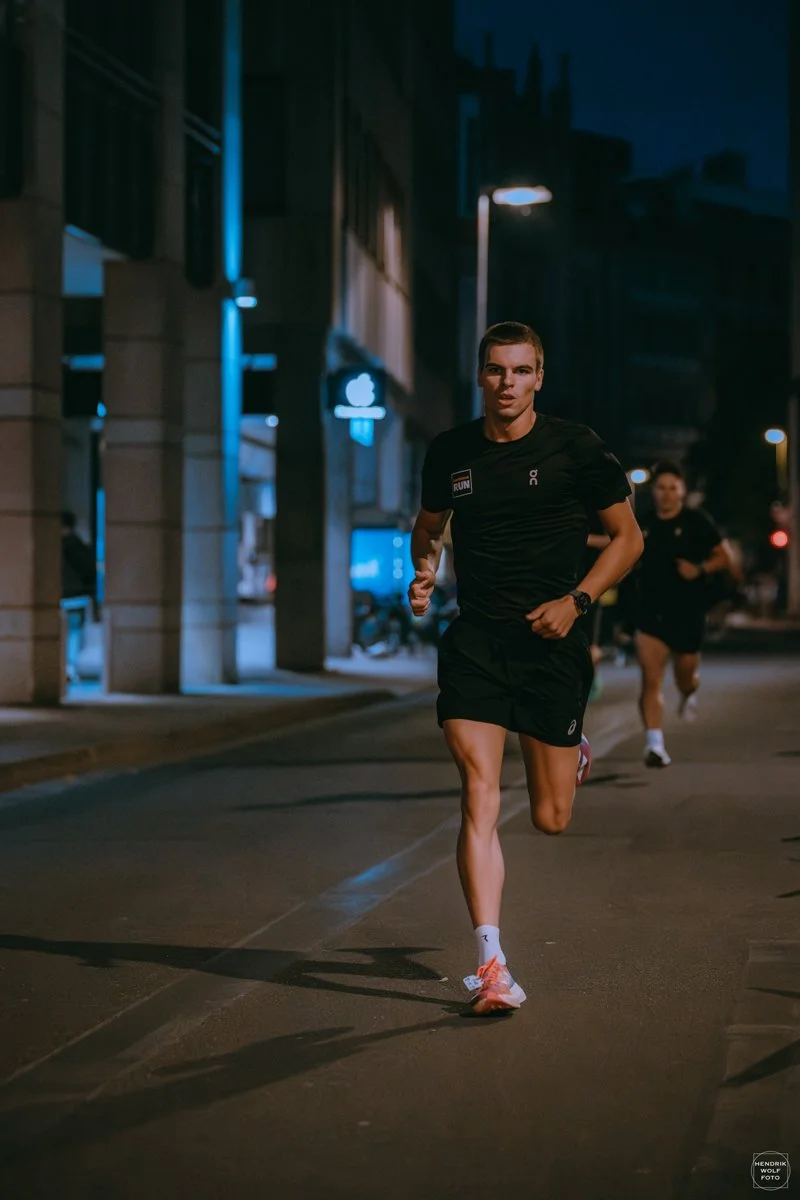 A young man running at night on a city street, wearing black athletic shorts, a black t-shirt, and pink running shoes.