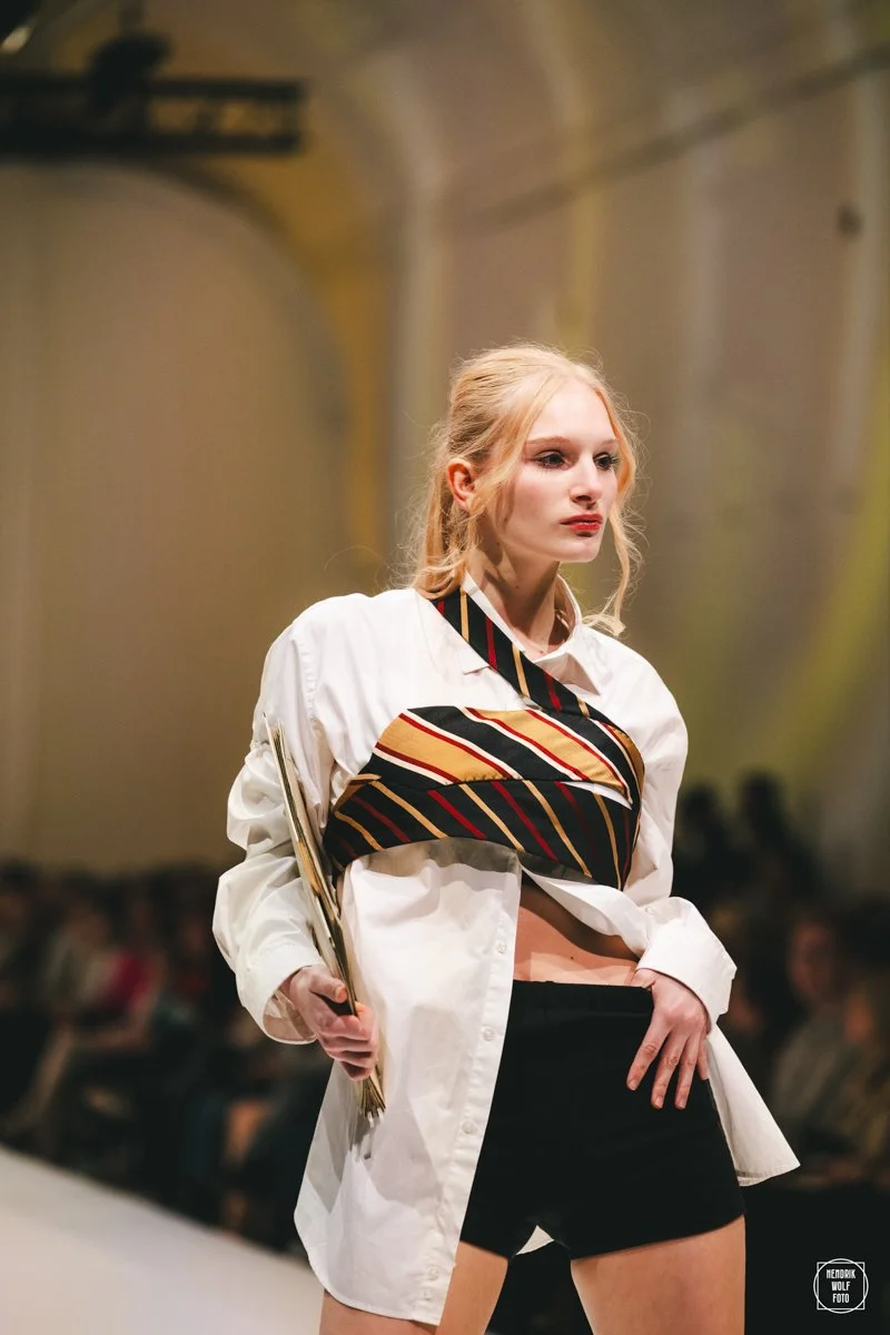 Fashion model walking on runway in a white shirt, black shorts, and a striped scarf across her chest, with a serious expression and holding a fan, in an indoor venue.