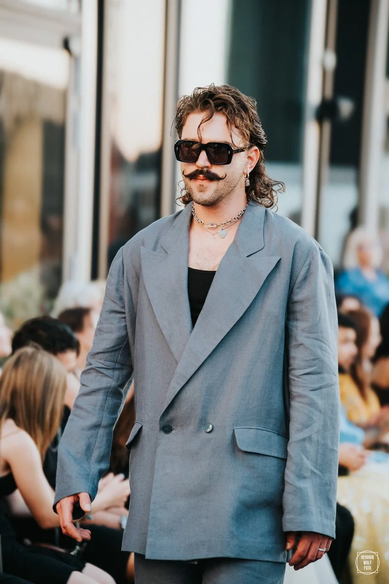A man wearing sunglasses, a grey suit, and jewelry, walking outdoors during the daytime with blurred people in the background.