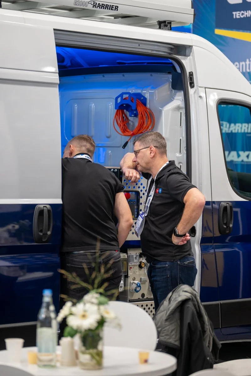 Two men inspecting equipment inside a service van at a trade show or exhibition. The van has open doors, and the men are focused on the equipment inside. The scene includes a table with a flower arrangement and disposable cups in the foreground.