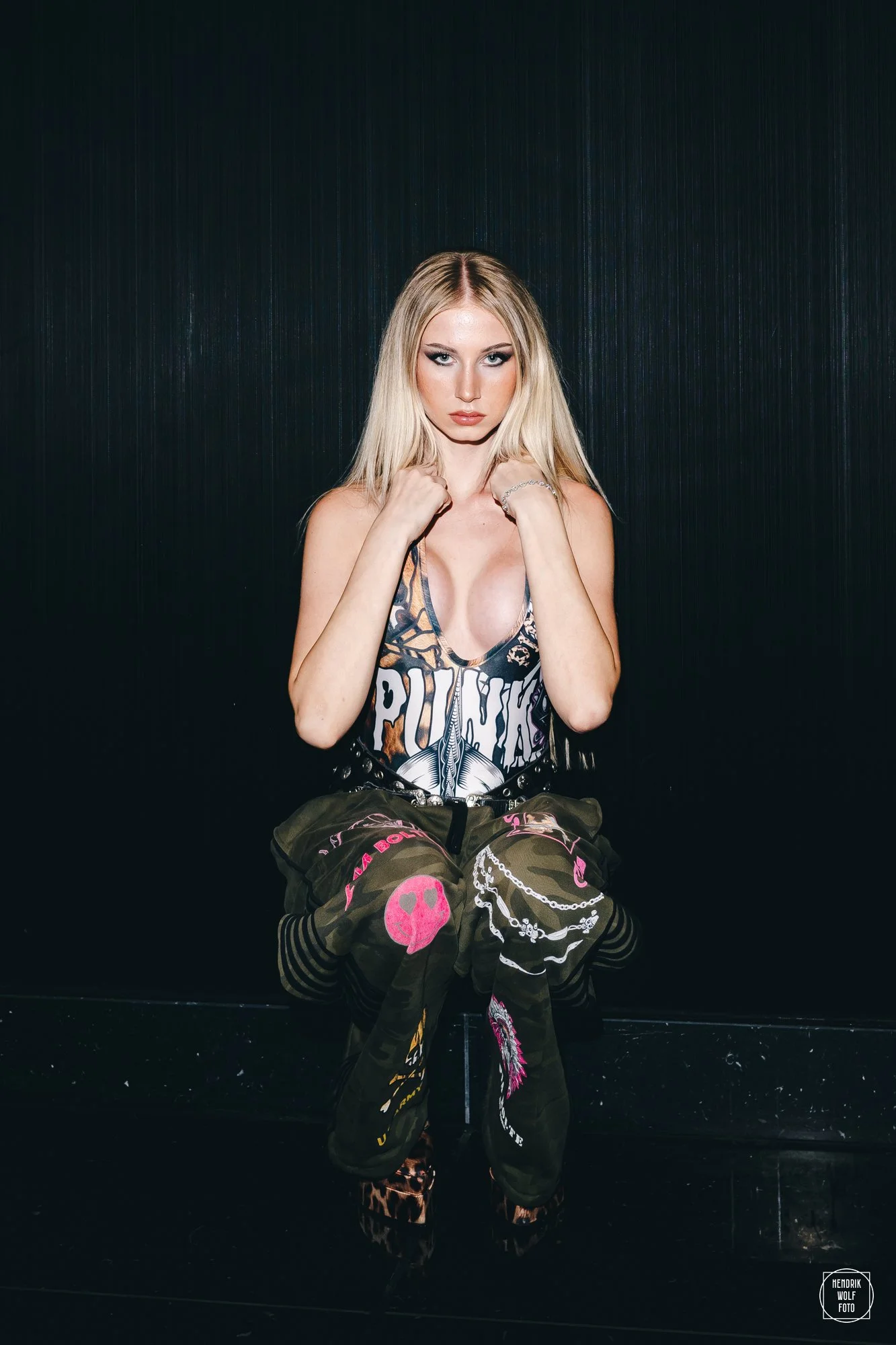 Young woman with long blonde hair and intense makeup, sitting against a dark textured wall, wearing a graphic tank top and baggy pants with pink skull and chain designs, and leopard print boots.