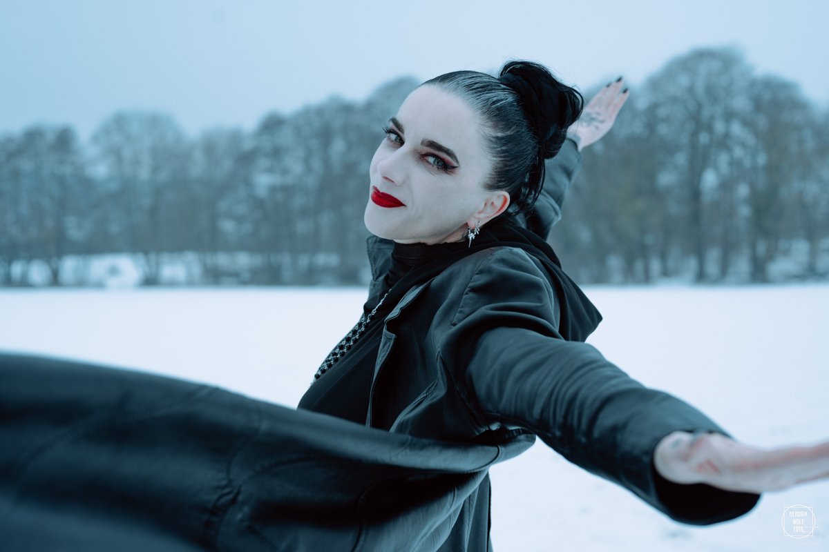 A woman with black hair in a bun, red lipstick, and makeup, wearing a black leather jacket, standing outdoors in a snowy landscape with trees in the background, extending her arm.