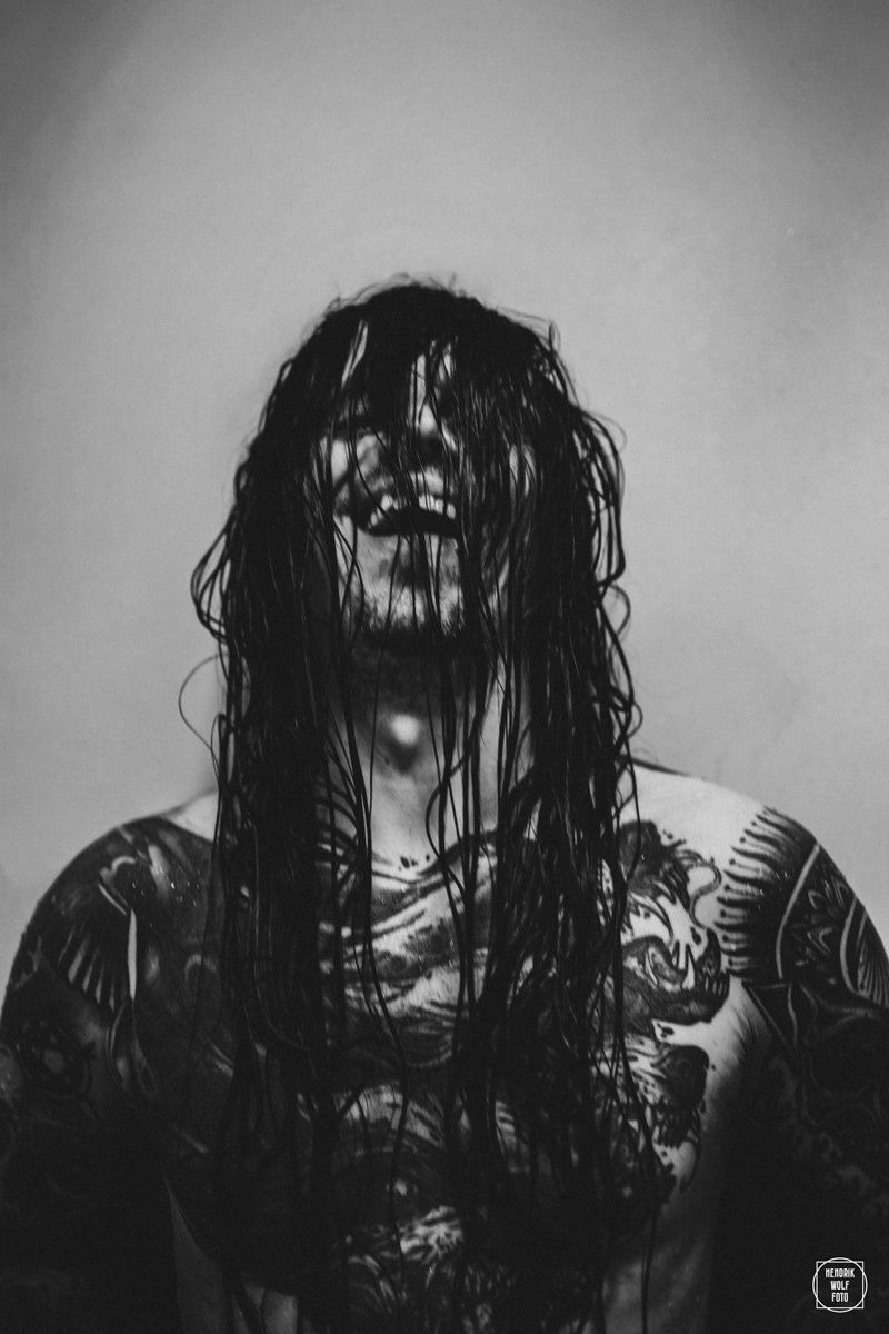 A person with wet, long dark hair covering their face, laughing, showing teeth and tattoos on shoulders and arms, in black and white portrait photograph.
