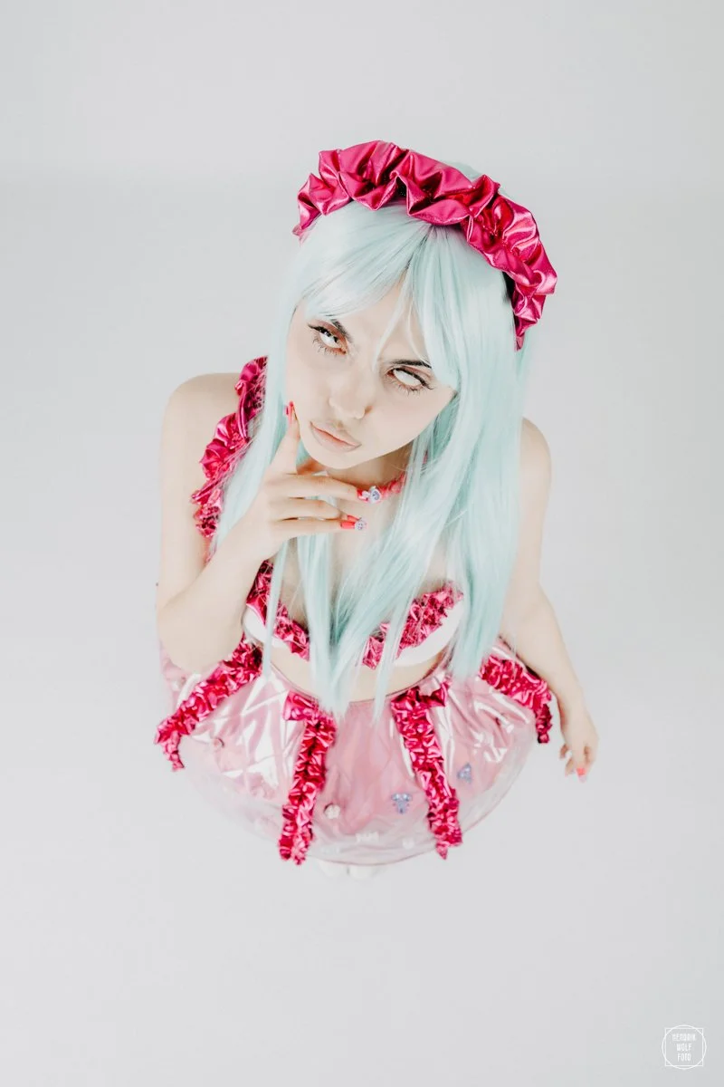 Person with long light blue hair wearing pink ruffled dress and matching headband, looking up with hand near mouth, posing against a plain white background.