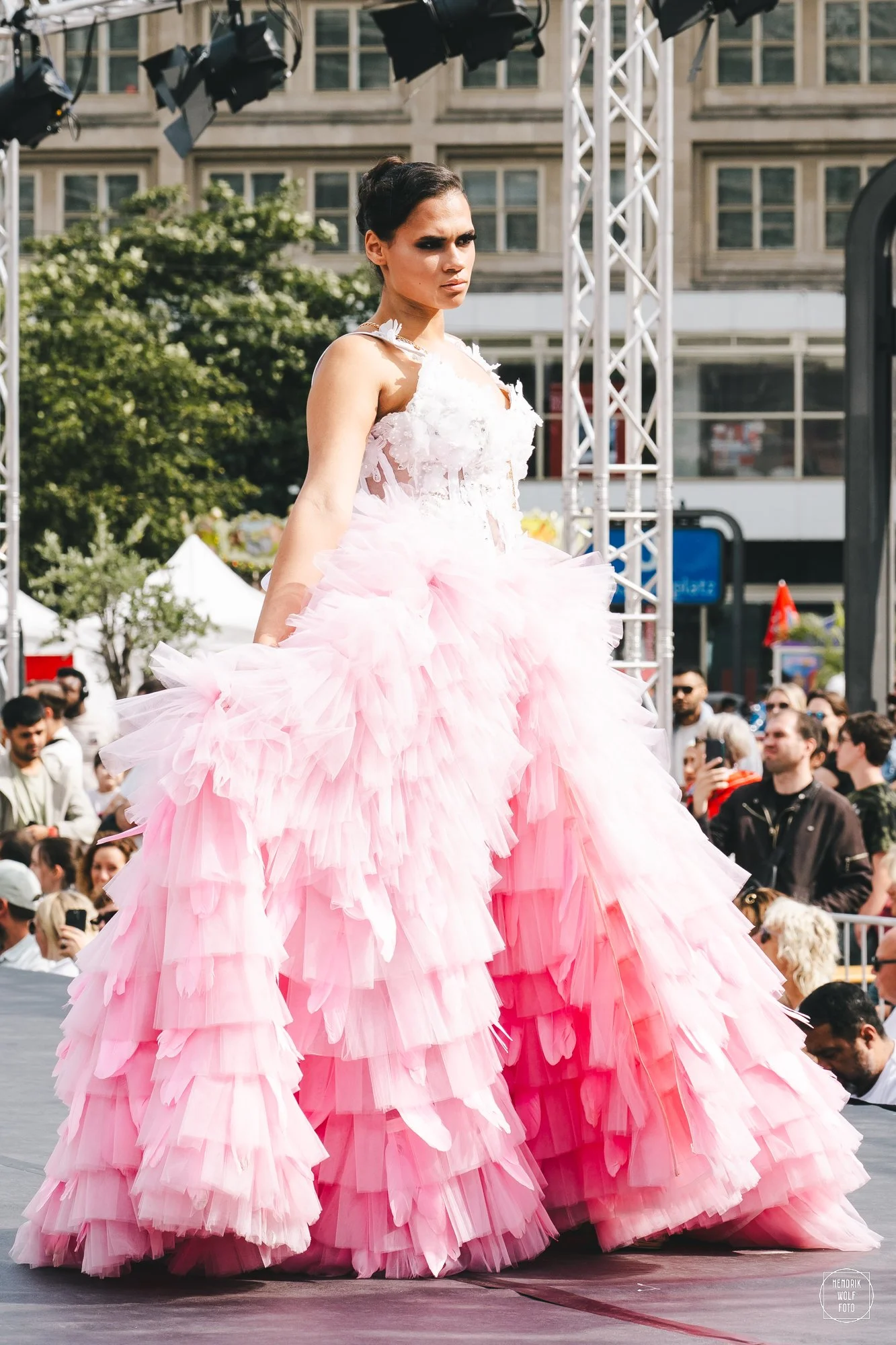Woman walking on stage in a large, pink, ruffled dress at an outdoor event, with onlookers and equipment visible in the background.