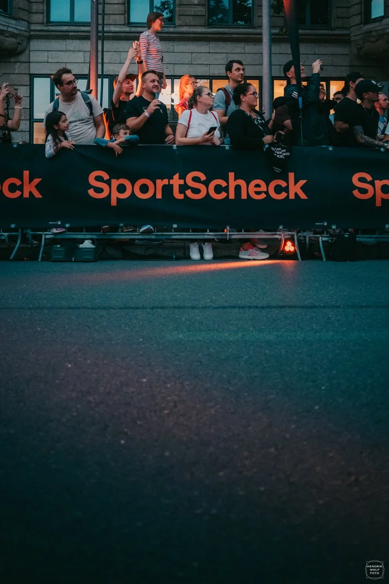 A group of people standing behind a black barrier with the red logo 'SportScheck' printed on it. Some are taking photos, and one person is speaking into a microphone. The scene appears to take place on a city street during an outdoor event at dusk.