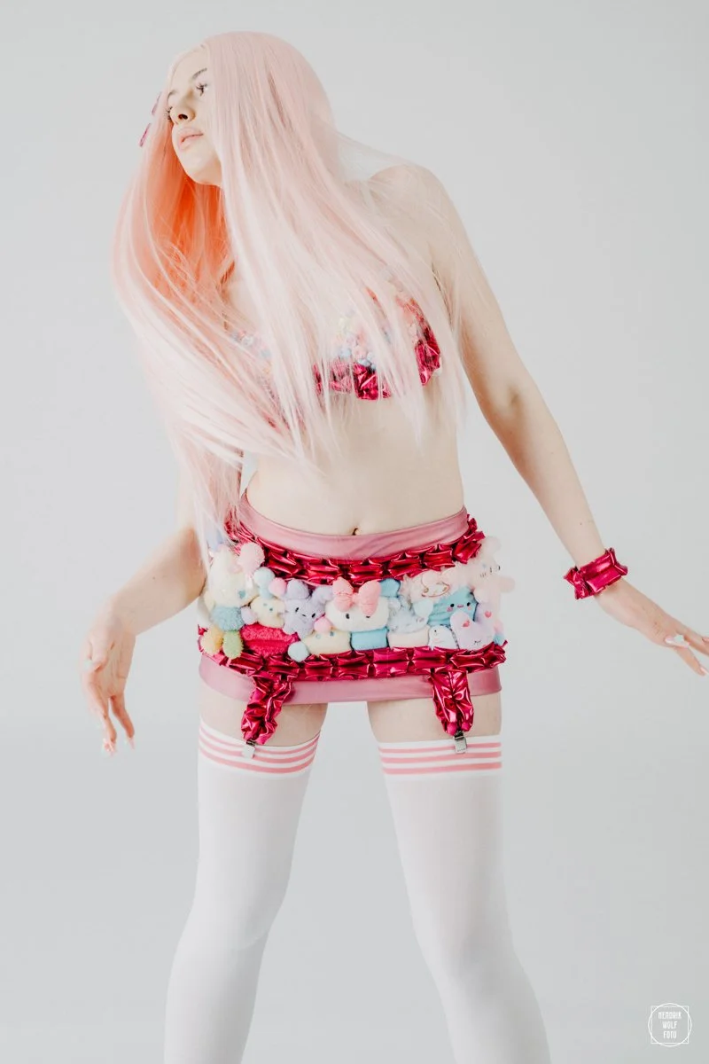 Young woman with long pink hair wearing a colorful, playful outfit with pastel stuffed animals, pink ribbons, and white thigh-high stockings with pink stripes.