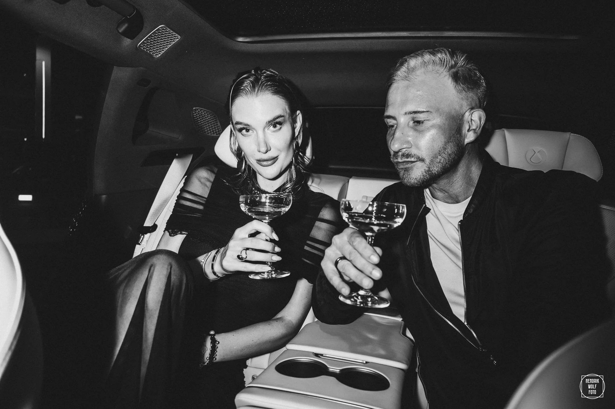 A black and white photo of a woman and a man sitting in the back of a luxury vehicle at night, holding cocktail glasses, possibly celebrating.