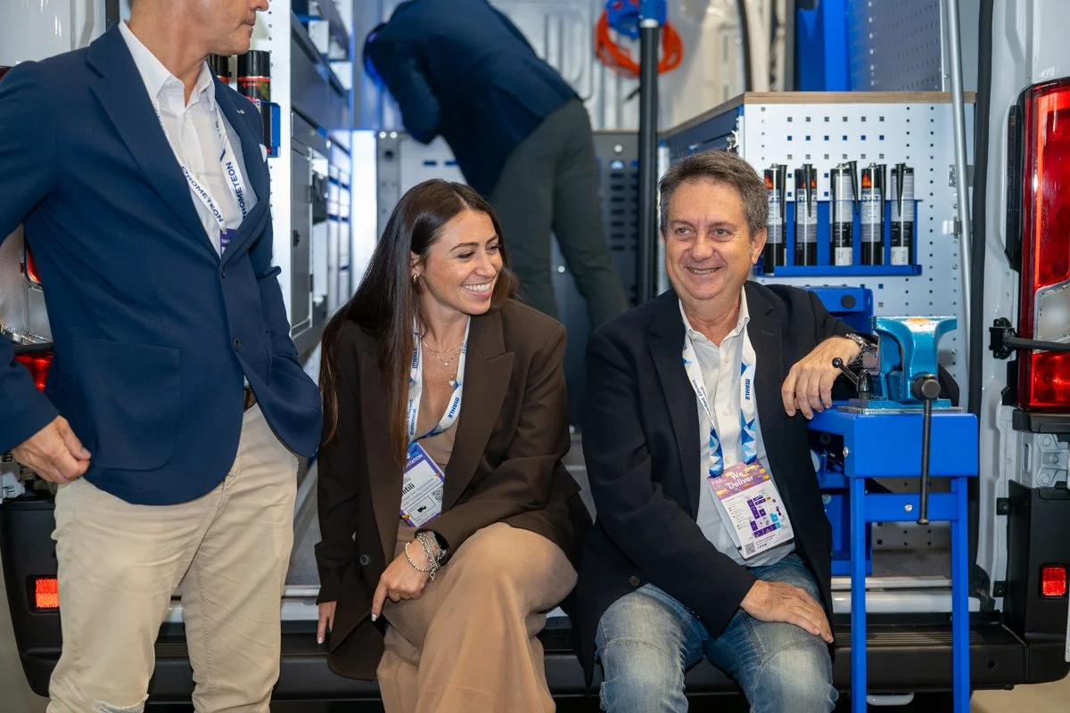 Three people in business attire smiling and talking near a work van with tools. Two are sitting and one is standing, with a woman in a brown blazer and a man in a dark blazer seated, and a man in a navy blazer standing.