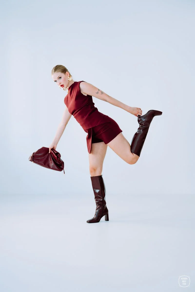A woman in a burgundy outfit and high-heeled boots poses with one leg raised, holding a matching clutch in a studio with a plain light background.