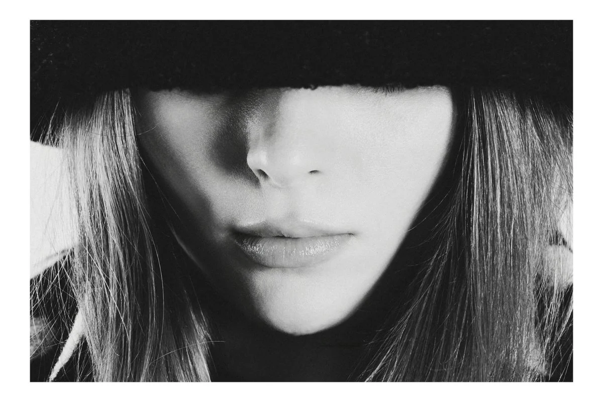 Close-up black and white photo of a woman with long hair wearing a wide-brimmed hat covering her eyes.