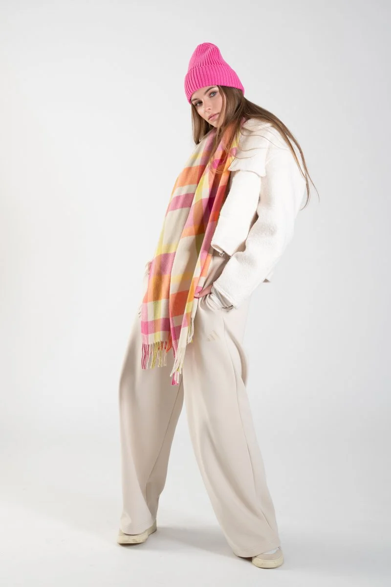 Woman wearing a pink knit beanie, white sweater, beige wide-leg pants, and a colorful plaid scarf, standing against a plain light gray background.
