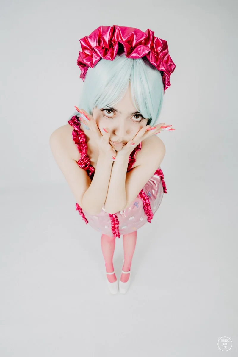 A woman with pastel blue hair wearing a shiny pink ruffled headband and dress, posing with her hands framing her face, standing against a plain white background.