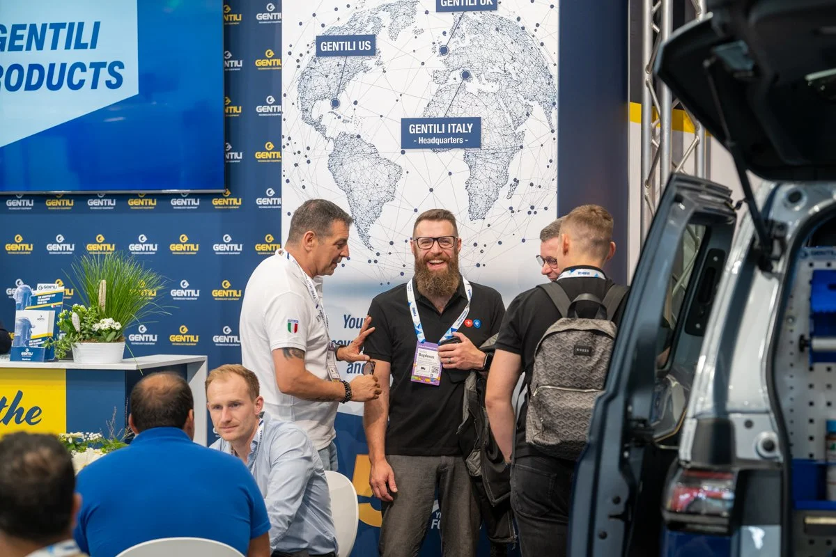 Group of men talking at a booth with banners that say 'GENTILI'. One man in the center is smiling and laughing, wearing glasses and carrying a backpack. A large world map displays the company's global offices, and there is a partially visible vehicle