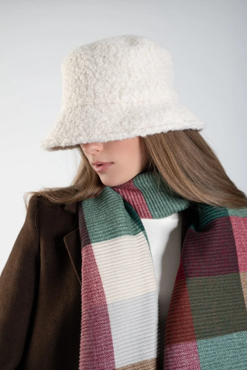 Woman wearing a beige fuzzy bucket hat, a brown coat, and a multicolored plaid scarf.