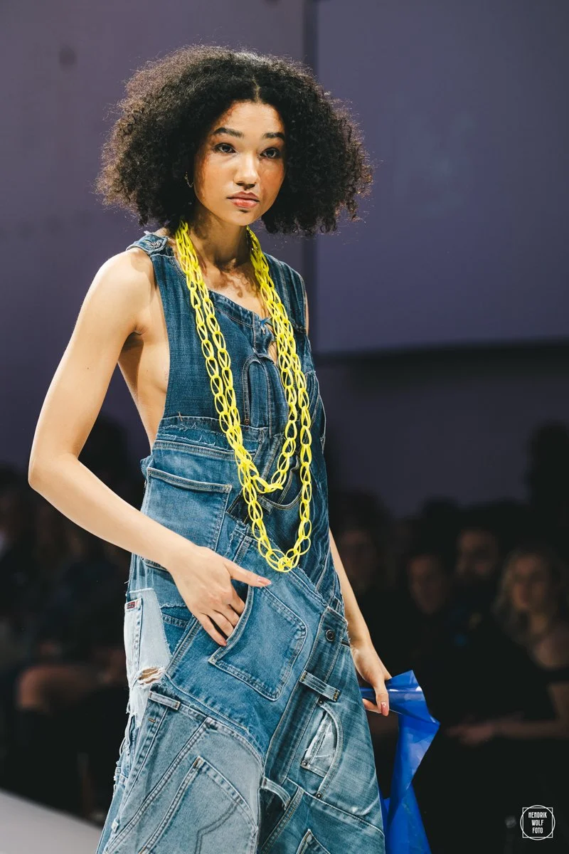 Female model walking on runway wearing sleeveless denim dress with large front pocket, yellow chain necklace, and holding blue fabric, with an audience in the background.