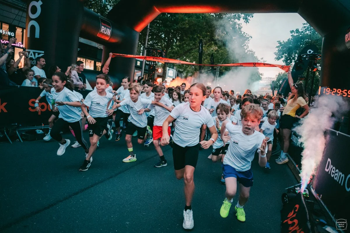 Children in athletic clothing start a race at the starting line under an inflatable archway with a digital timer in an outdoor setting surrounded by trees and spectators.