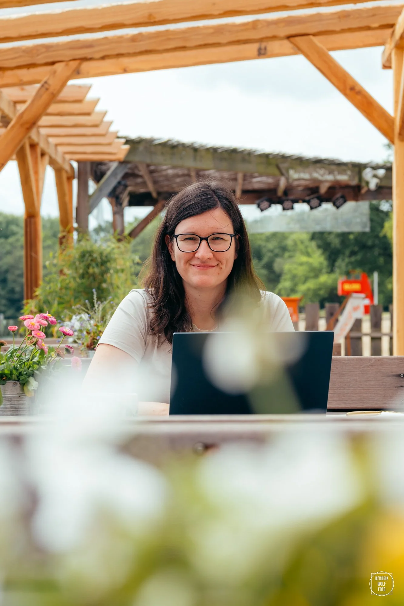 A young woman with glasses and dark hair, smiling while sitting outdoors at a table with a laptop, surrounded by colorful flowers, with a wooden pergola structure above her and lush greenery in the background.