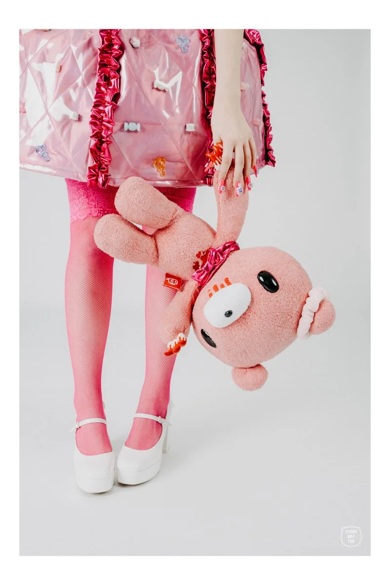 A person dressed in pink, wearing high heels, pink fishnet tights, and a pink shiny jacket, holding a pink stuffed teddy bear upside down by its arm.