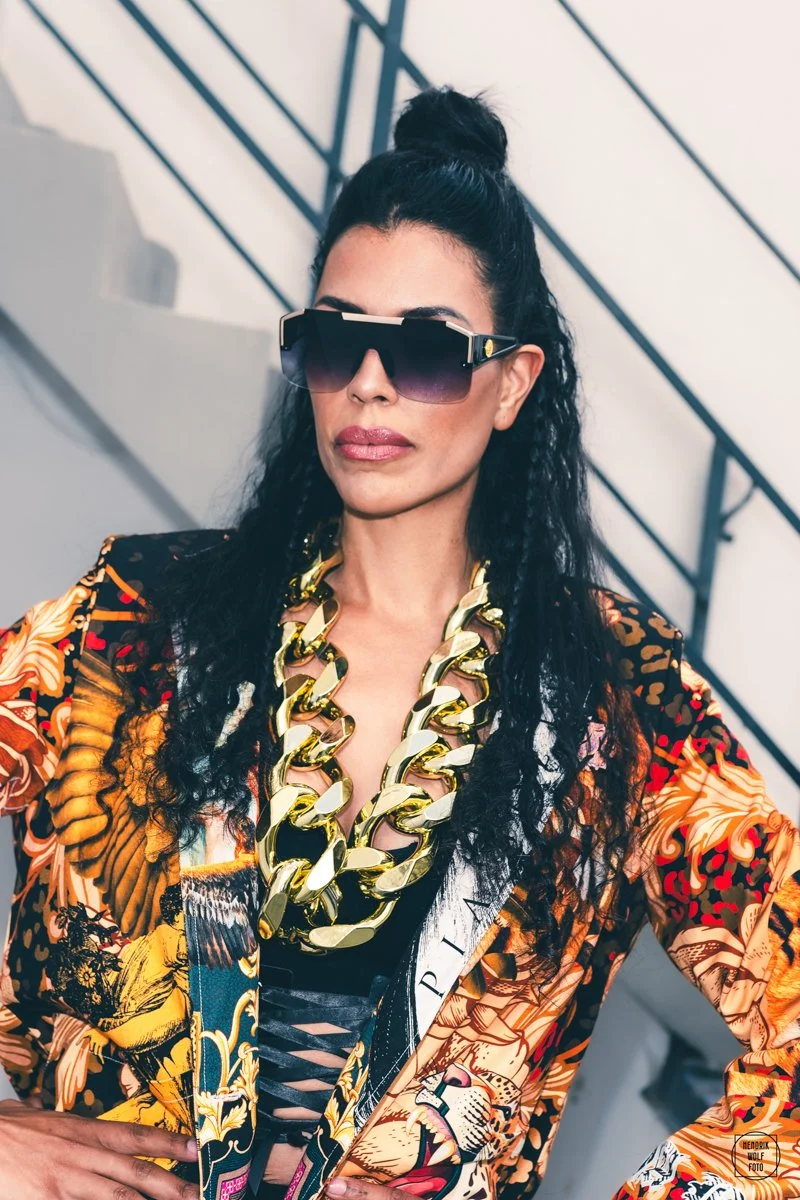 A woman with dark, curly hair in a high bun wearing large, fashionable sunglasses, a thick gold chain necklace, a colorful patterned blazer with floral and bird motifs, and a black lace-up top underneath. She is standing indoors against a staircase with black railing.
