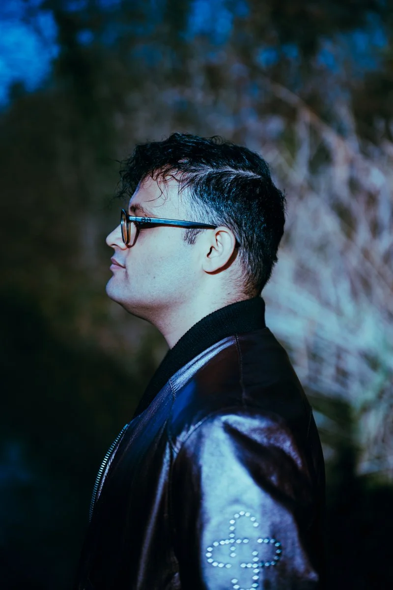 Profile of a man with glasses, short dark curly hair, wearing a black leather jacket with a clover design made of rhinestones on the sleeve, standing outdoors during dusk.