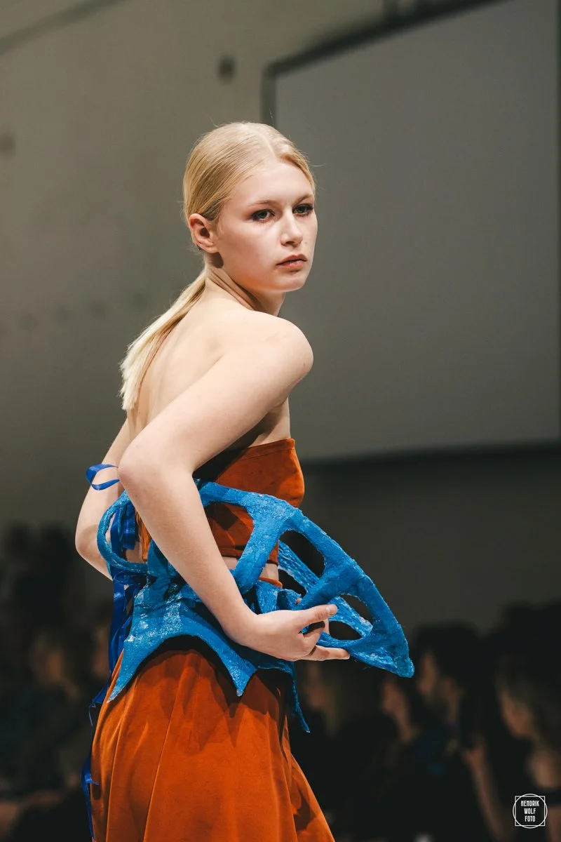 A female model with blonde hair styled in a ponytail walking on a runway at a fashion show.