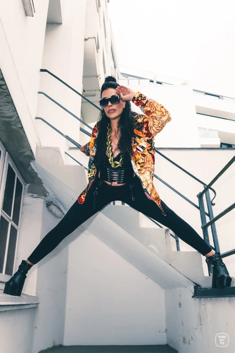 A woman in black pants, black high-heeled shoes, and a colorful patterned jacket poses on outdoor stairs. She is wearing sunglasses and has her hair styled in a high bun, with one hand saluting and the other resting on the railing.