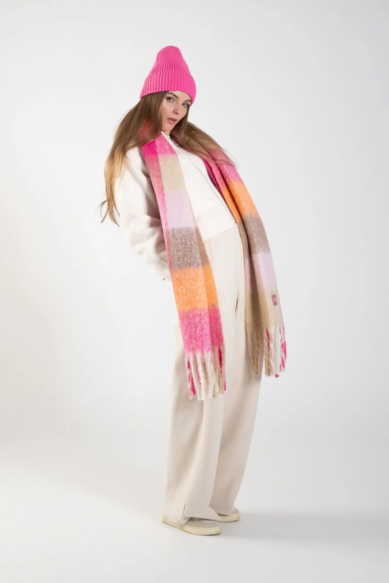 Woman in pink knit hat, white outfit, and multicolored scarf posing against a plain background.