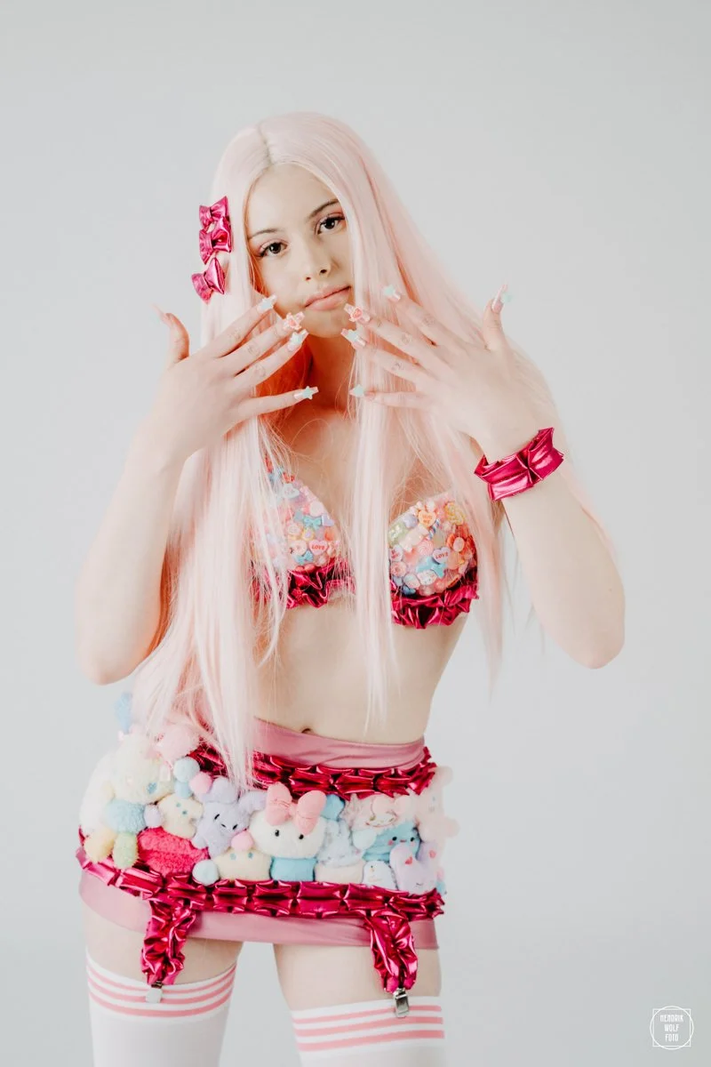 Young woman with long pink hair wearing colorful, stuffed animal-themed lingerie, pink bows in her hair, and striped thigh-high stockings, posing with her hands near her face against a plain light background.