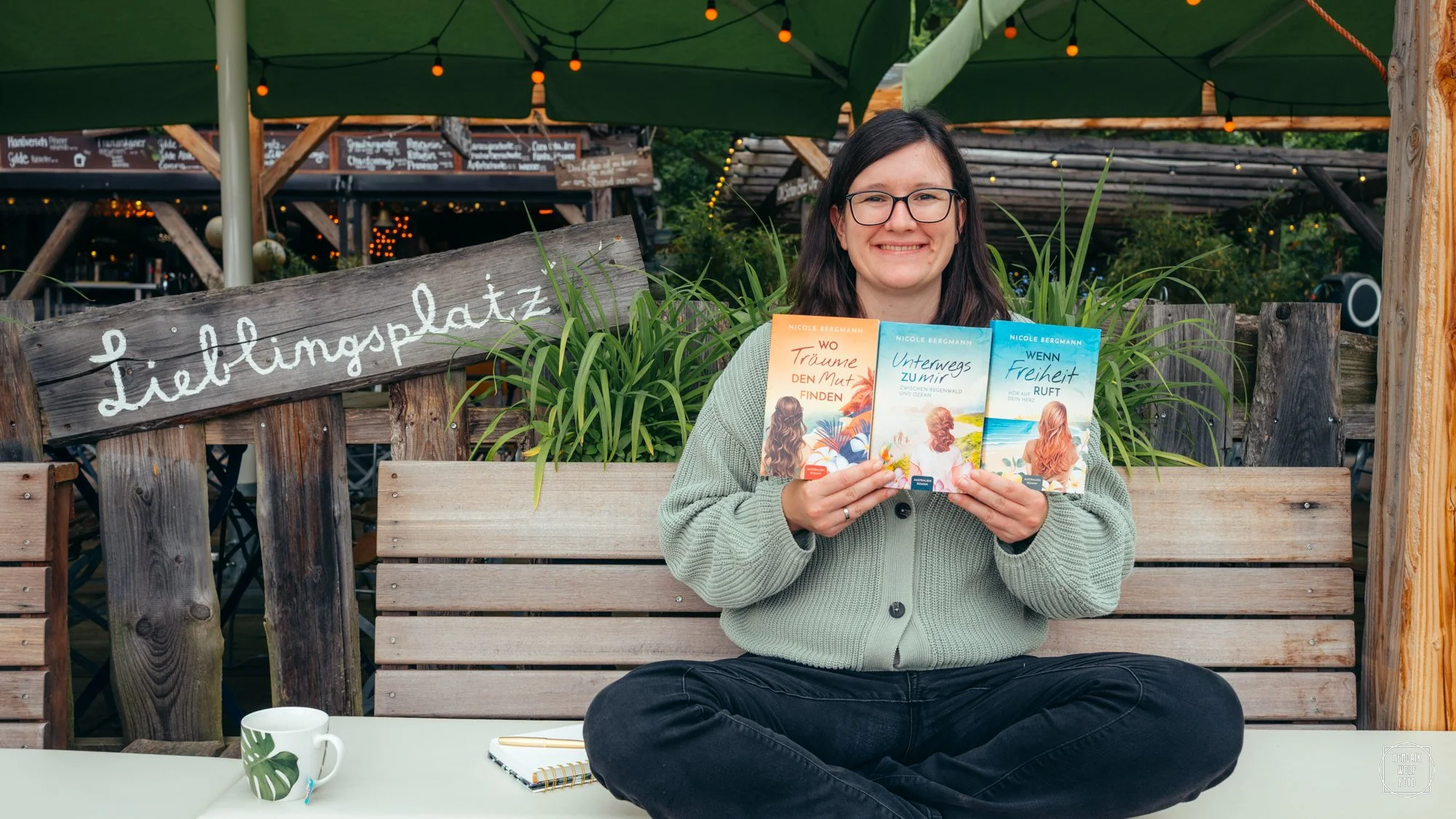 A woman with glasses and dark hair, dressed in a gray sweater, is sitting cross-legged on a bench outdoors, holding three books with colorful covers. She is smiling. Behind her, there is a wooden fence with a sign that reads 'Lieblingsplatz'. The background includes string lights and greenery, suggesting a cozy outdoor setting.