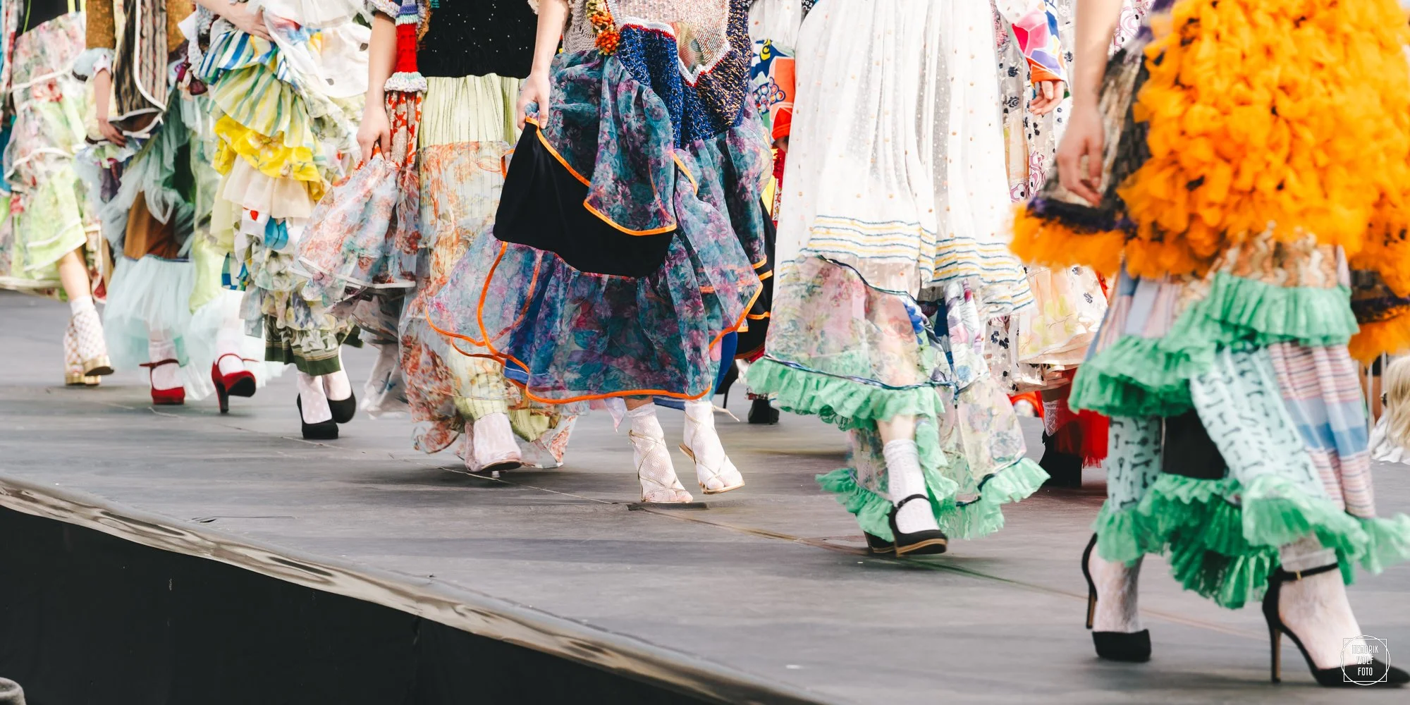 A group of women walking on a stage, wearing colorful and elaborate skirts and tops, possibly performing or participating in a cultural event or fashion show.