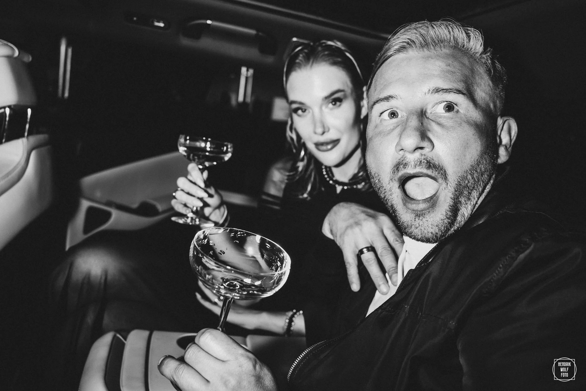 Black and white photo of a man and woman in a car, holding glasses, with surprised expressions, woman looking at the camera, man with mouth open, in a vintage style setting.