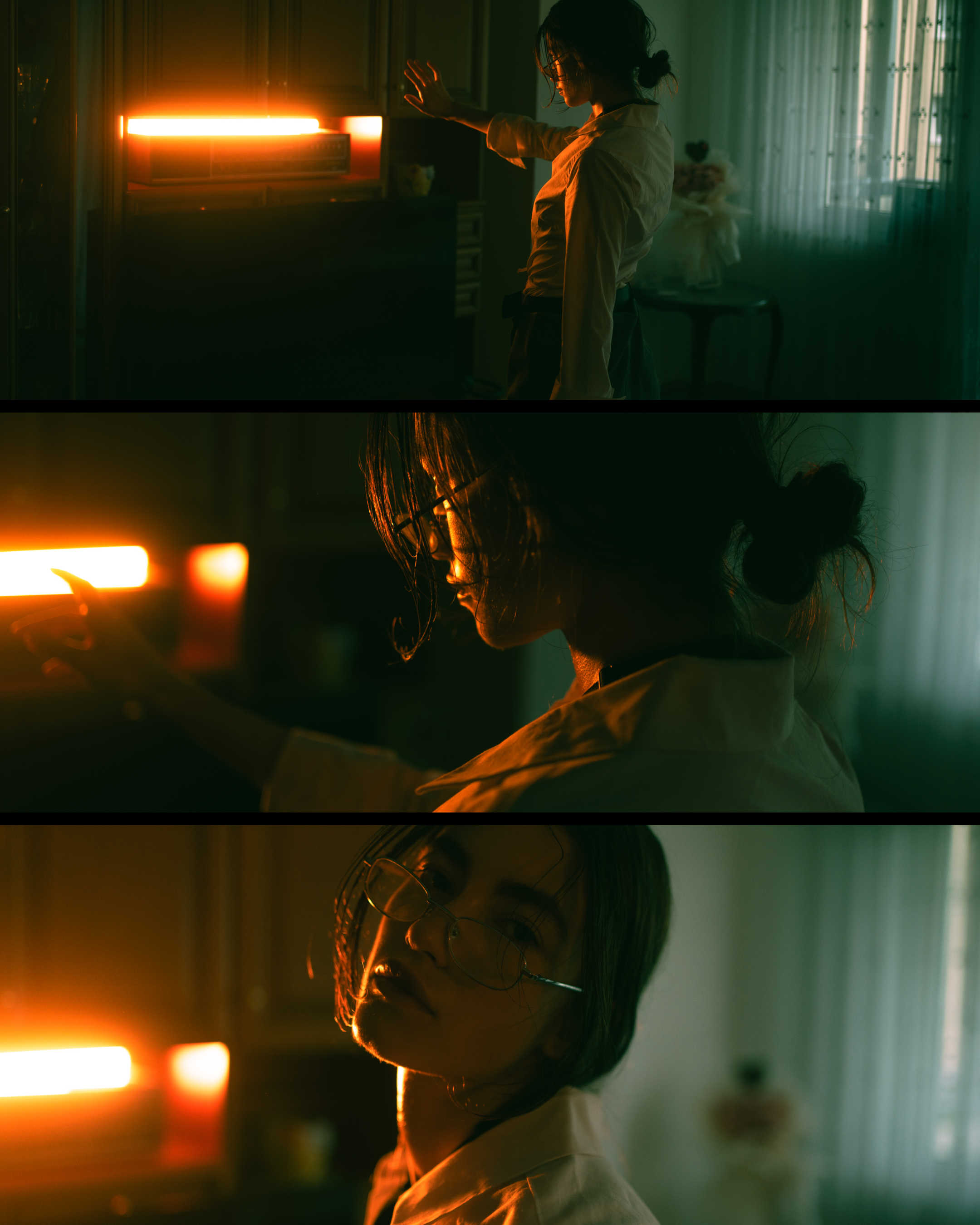 A woman with curly hair and glasses in a white shirt and apron stands in a dimly lit room with greenish light, facing a bright orange glowing light source, possibly a heater or lamp, with her hand raised towards it.