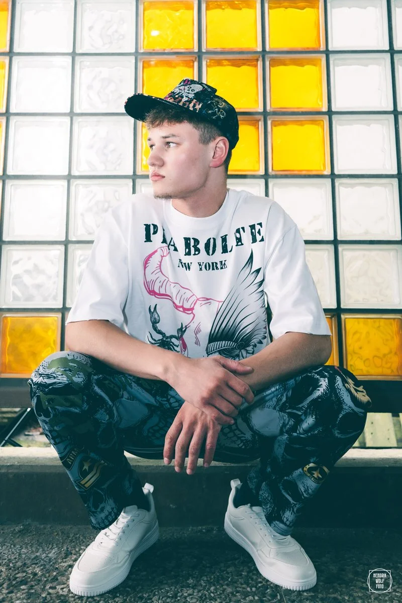 A young man is sitting on the ground in front of a wall made of glass blocks, some of which are yellow. He is wearing a white T-shirt with a graphic design and text, patterned pants, white sneakers, and a cap. He is looking to his left with a thoughtful expression.