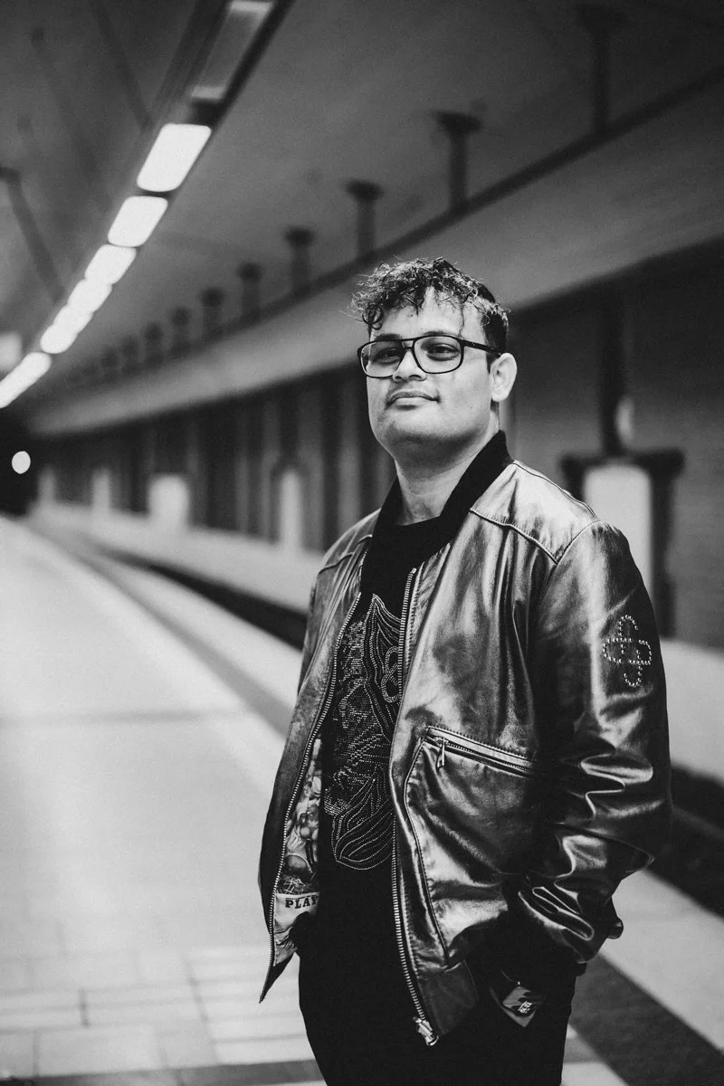 A man with glasses and curly hair wearing a leather jacket standing on a subway or train platform.