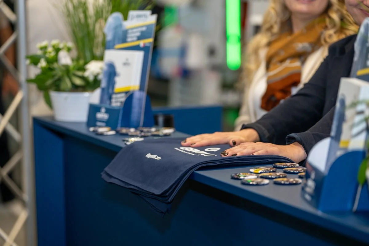 A person is resting their hands on a table display at a promotional booth, with buttons, bags, and brochures arranged on the table. A woman with blonde hair, wearing a patterned scarf, is visible in the background.