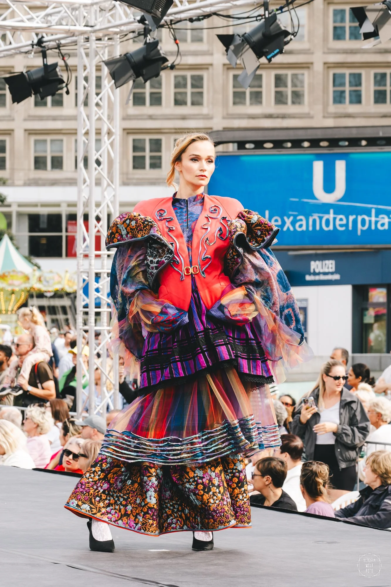 A woman on a runway wears a colorful, layered outfit with floral and striped patterns, and a pink jacket with embroidered details. She stands in front of a crowd, with festival booths and a blue sign in the background.
