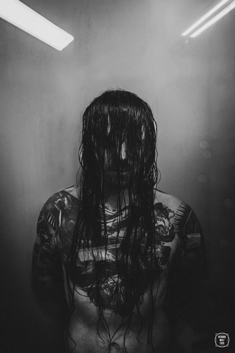 A person with long wet hair covering their face, showing tattoos on arms and shoulders, standing indoors with a plain wall and fluorescent lights above.