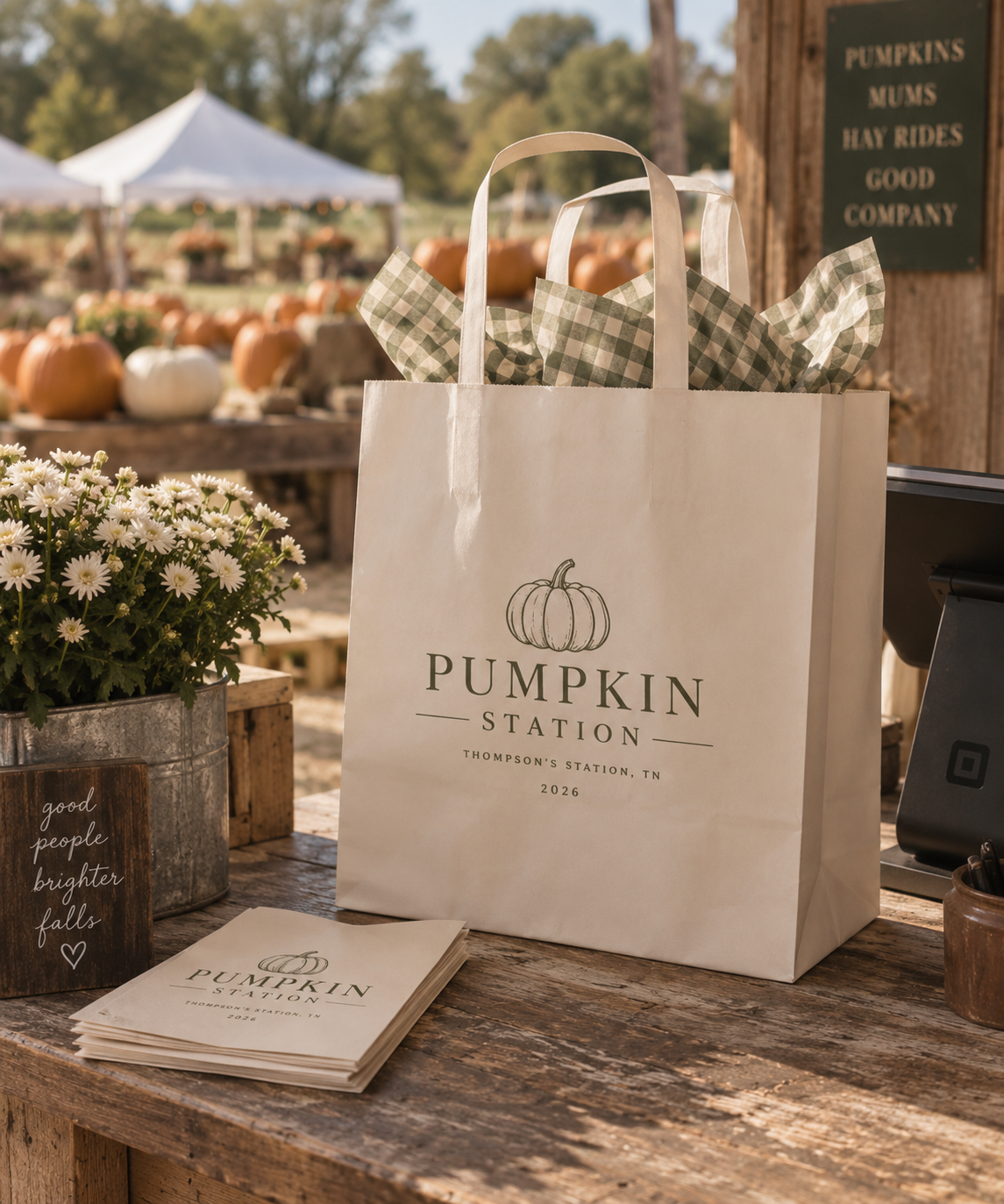 A white paper bag with a pumpkin illustration and label reading "Pumpkin Station" on a rustic wooden table. In the background, pumpkins of various colors and sizes are displayed outdoors, along with a tent and trees. There are also some white flowers and a small sign that says "good people brighter falls."